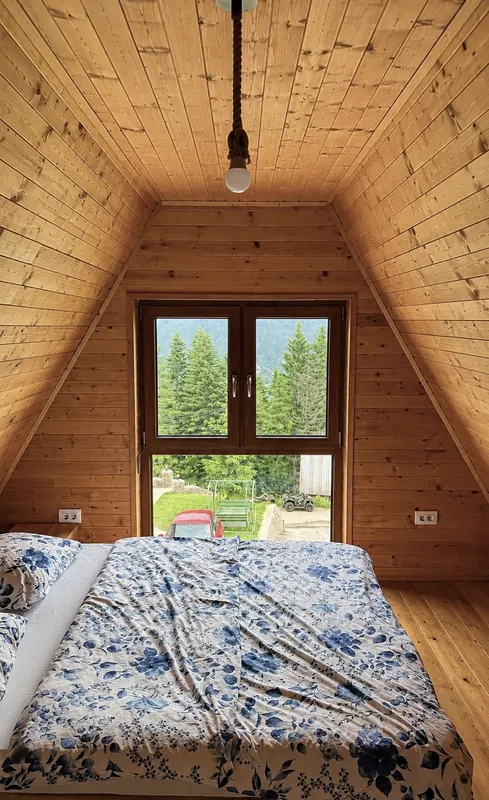 Bedroom under the sloping pine roof with a window onto spruce trees